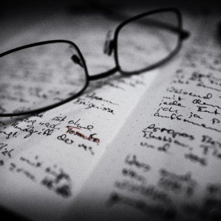 Schwarz-weiß Bild vom inneren eines Notizbuchs in dem beide Seiten voller handschriftlicher Notizen sind. Eine Brille liegt auf der linken Seite, ein Wort scheint rot nachgezeichnet zu sein: Toronto.