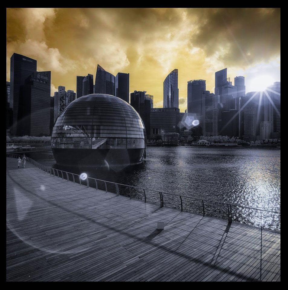 Infrarodshot taken in Singapore. Sky is yellow, much of the picture is grey-blue. There is a skyline in the back, a bay in the middle and a wooden walkway in the front. At the left middle there's a sphere in the water (it houses Singapore's flagship Apple Store). There's a lens flare and a sun star.