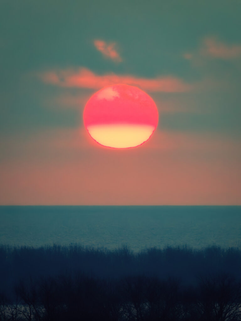 A stylized, post-processed version of the sunset with a cinematic, teal-and-orange color palette. The sky is a rich, moody teal, contrasting heavily with the sun, which now glows intensely with neon pink on top and bright yellow on the bottom. The image has a soft, dreamy quality with deep, matte shadows in the tree line.