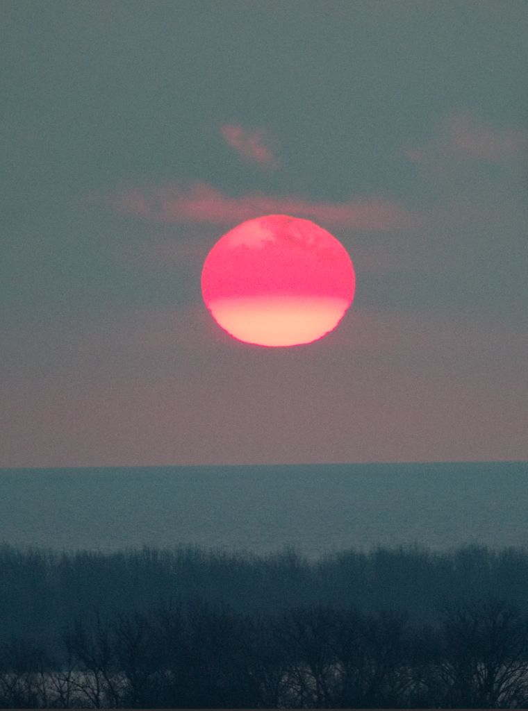 A raw, unprocessed photograph of a sunset over a body of water. The image has a muted, low-contrast look with a dusty mauve and grey sky. The sun is a large, soft orb, reddish on the top half and pale yellow on the bottom, hovering above a dark, silhouetted line of trees in the foreground.